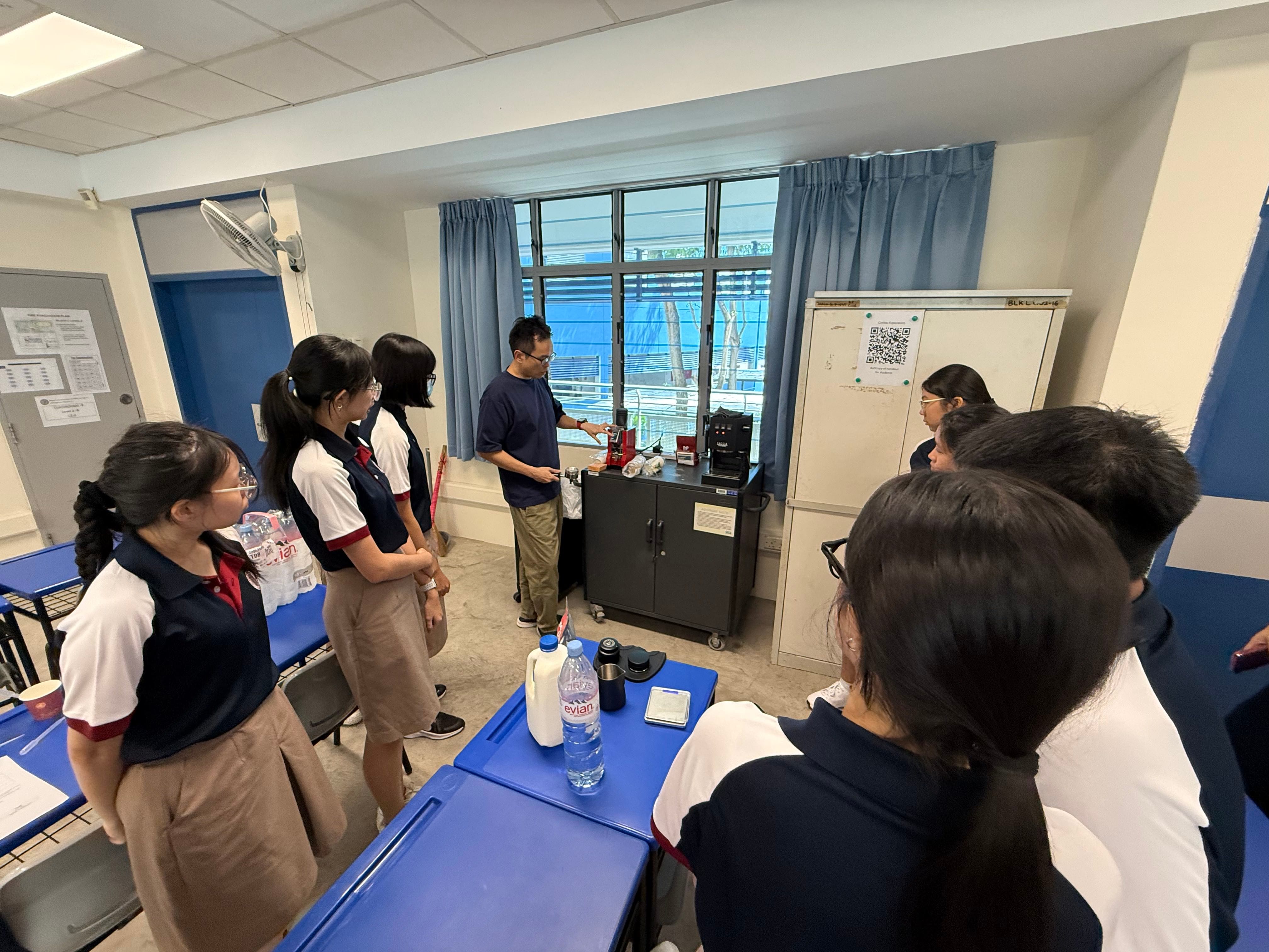 A group of students in navy blue polo shirts gather around a teacher who demonstrates coffee brewing using an espresso machine and grinder, with a bottle of milk and other equipment on the table nearby.